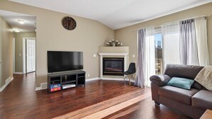 Uma sala de estar com piso de madeira, uma televisão de tela plana em um suporte, uma lareira e um sofá com almofadas.