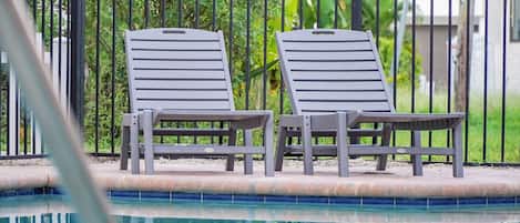Two grey lounge chairs by a swimming pool with a metal fence and greenery in the background.