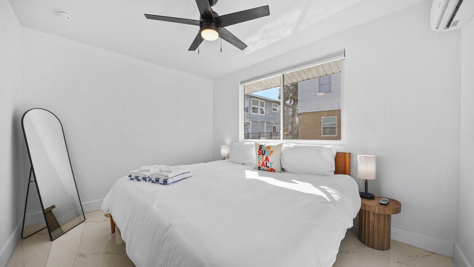 A bedroom with a bed, a ceiling fan, a nightstand with a lamp, and a window with a view of buildings.