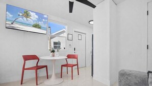 Ein modernes Zimmer mit weißer Wand, einem runden Tisch mit zwei roten Stühlen, einem grauen Sofa und einem Fernseher, der eine Strandansicht zeigt.