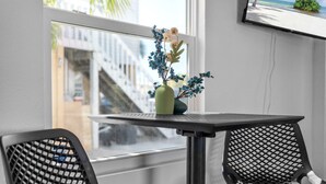 Une petite table ornée d’un vase de fleurs et deux chaises en osier sont placées devant une fenêtre donnant sur des immeubles.