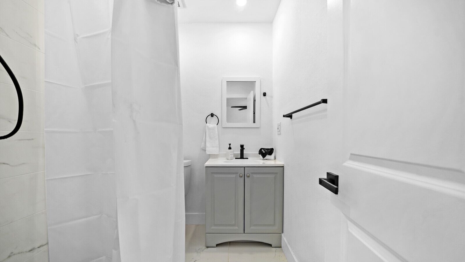 A narrow, white-walled bathroom with a toilet, sink, and mirror.