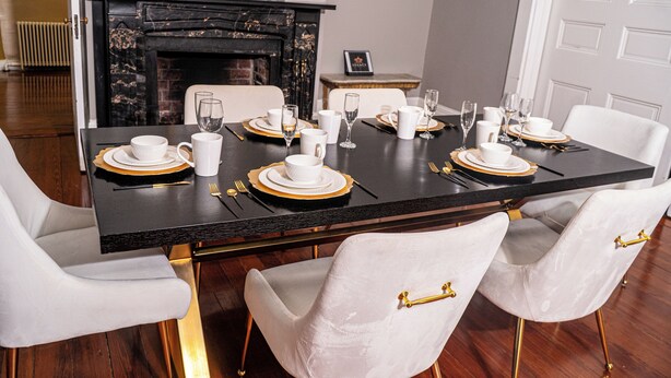 Una mesa de comedor con sillas blancas, detalles dorados y una chimenea al fondo.