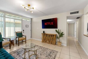 Una sala de estar con una televisión montada en la pared, un sofá, una silla, una mesita y una planta decorativa.