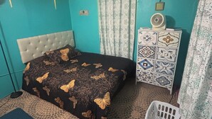 Een slaapkamer met een bed, een dressoir, een ventilator en een wasmand.