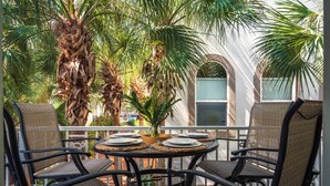 A balcony with a round table set for dining, surrounded by wicker chairs, palm trees, and a view of a building with arched windows. - Only 3 minutes walk to beach - Spacious Destin Home, Newly-renovated (Destin)