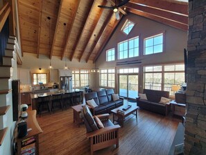 A spacious living area with a large couch, a coffee table, and a fireplace. There is a kitchen area with a dining table and chairs. The room has high ceilings with exposed wooden beams and large windows. - 4 Bed Luxury White Mtn Retreat w/ Stunning Views! (Campton, NH)