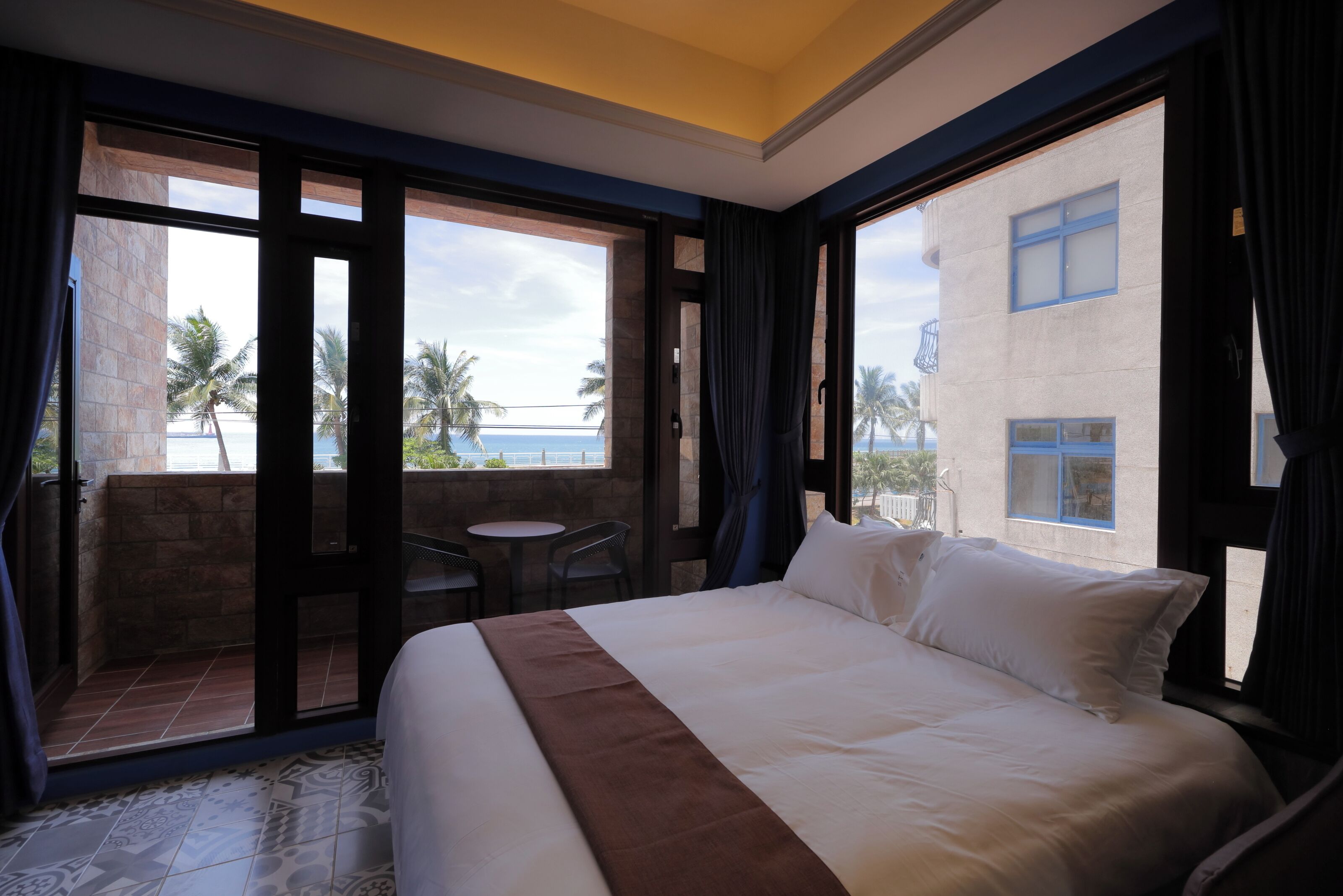 A hotel room with a large bed, a small table, and a chair. The room has a balcony with a view of palm trees and the ocean.