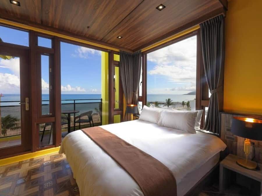 A hotel room with a large bed, a view of the ocean, and a balcony with a table and chairs.