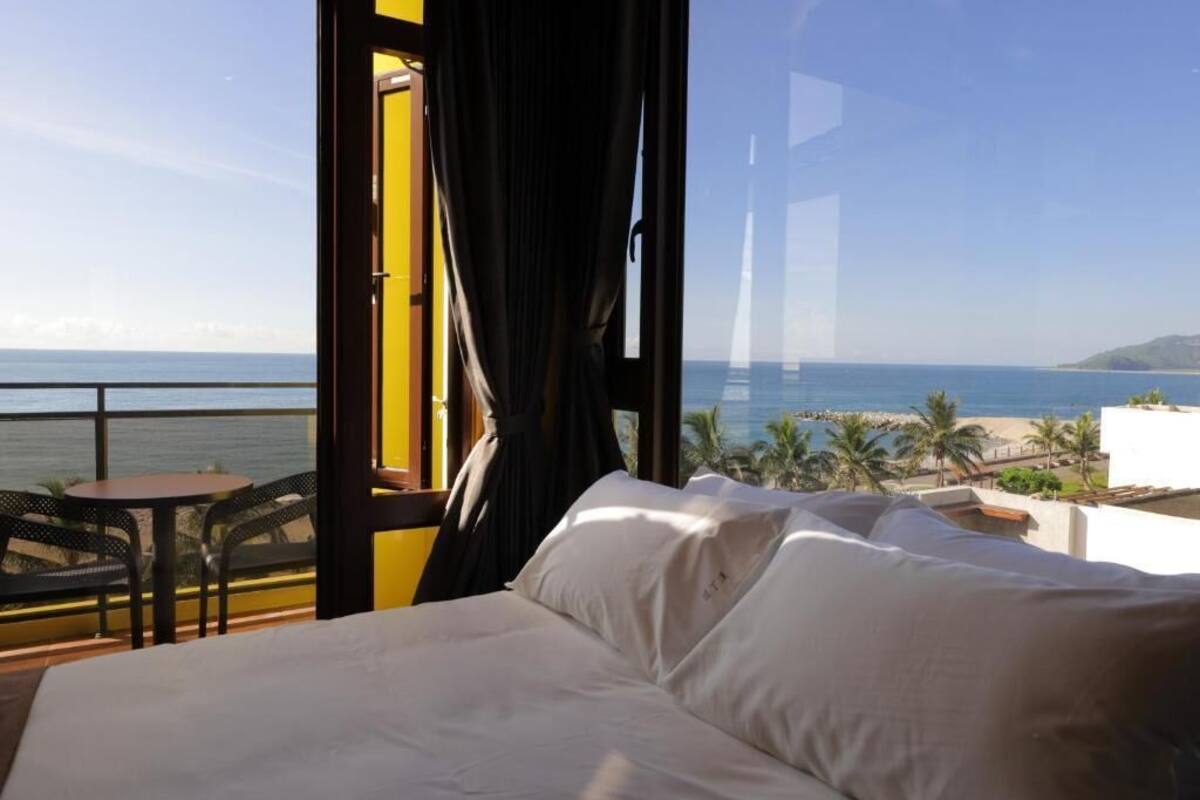 A hotel room with a bed, a small table, and a view of the ocean and beach.