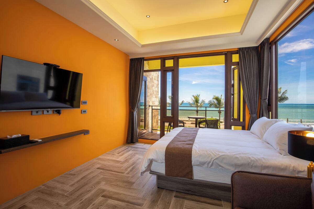 A hotel room with a large bed, a TV mounted on the wall, and a balcony with a view of the ocean.