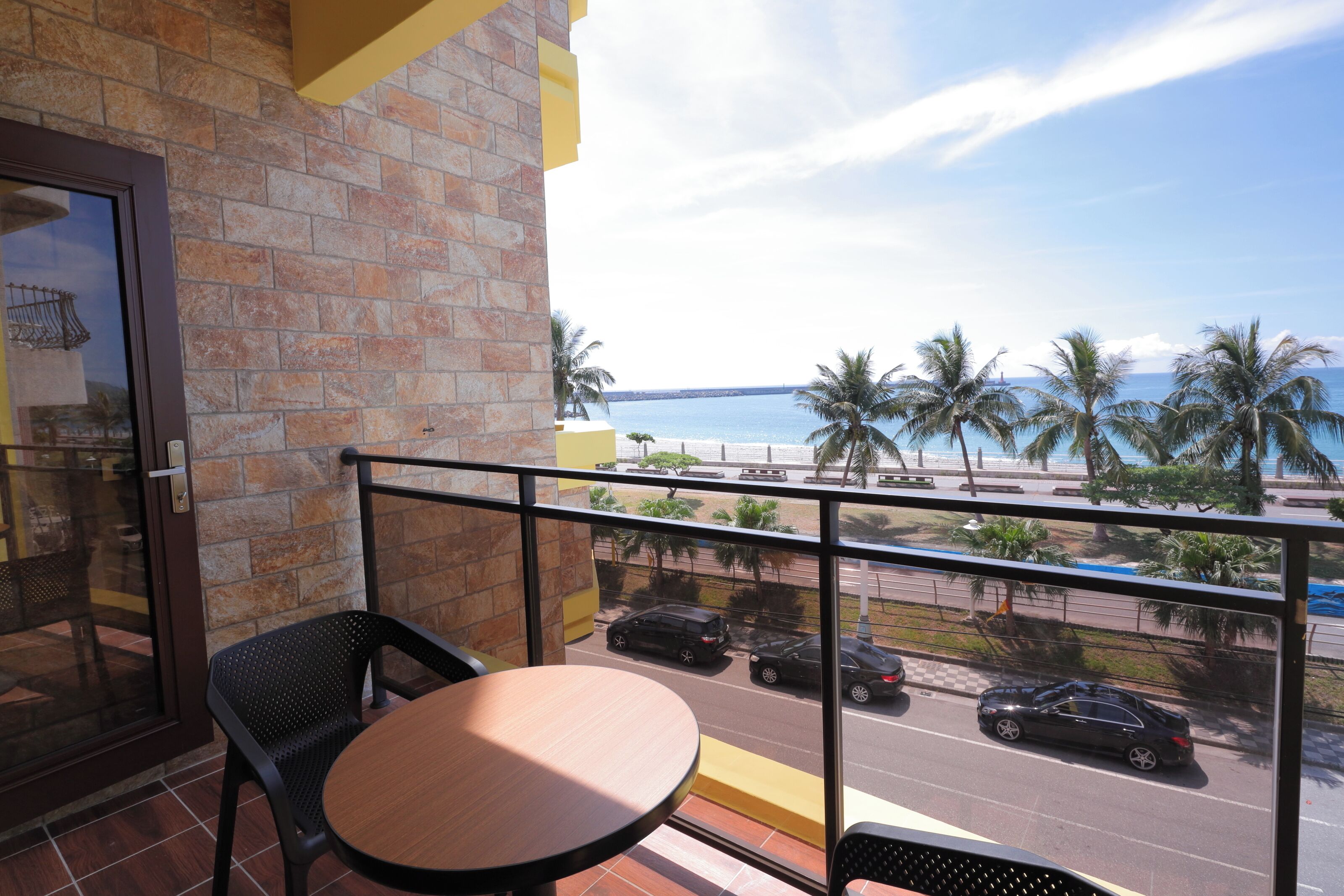 A balcony with a table and chairs, overlooking a beach and a road with cars.