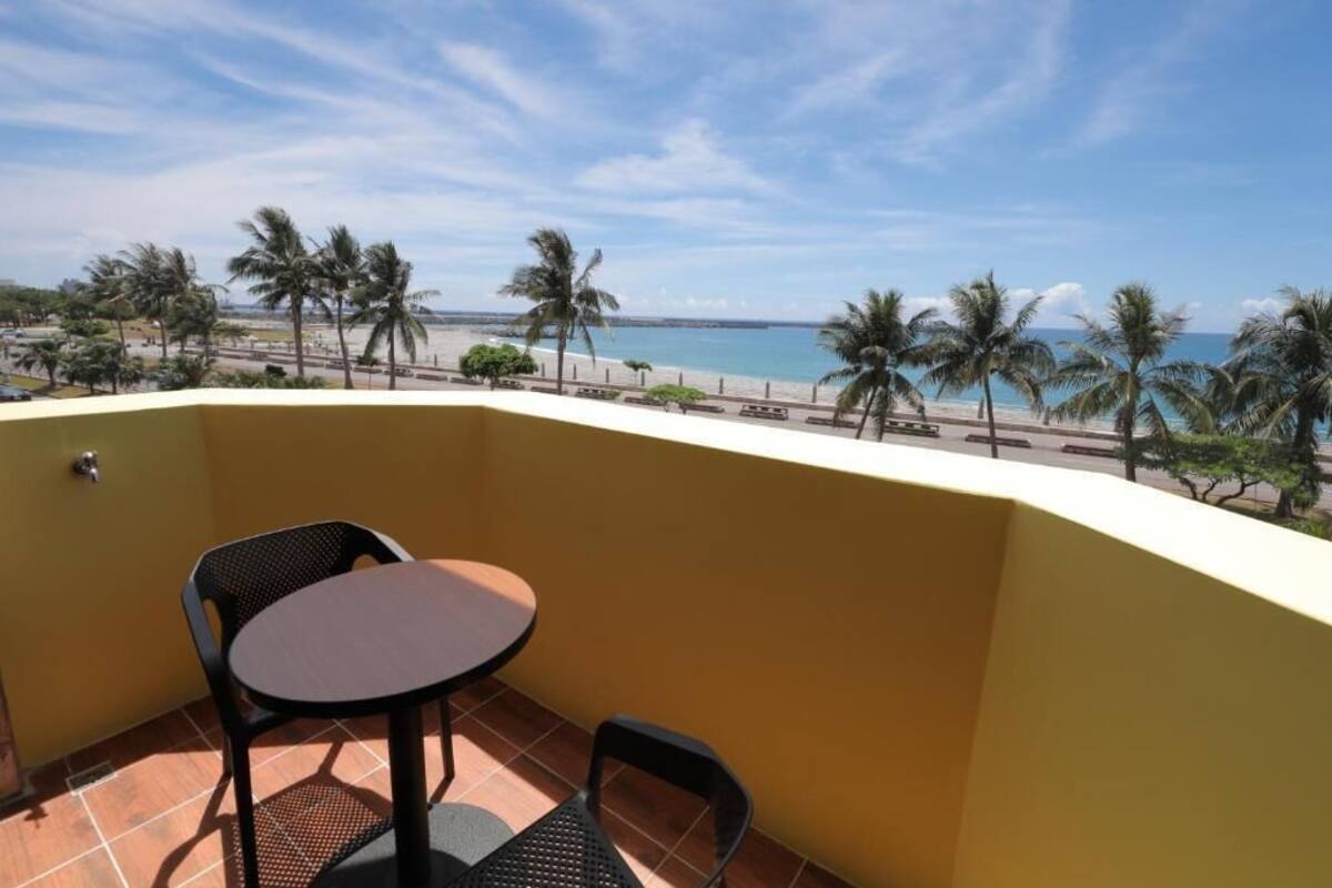 A balcony with a table and chairs, overlooking a beach with palm trees and a clear sky.