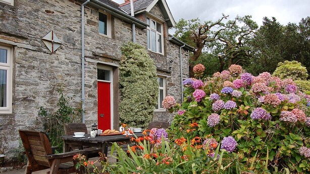 Una casa de pedra amb una porta vermella, un jardí amb hortènsies i una taula amb articles per esmorzar.