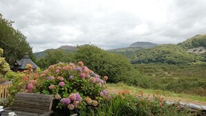 Un banc de fusta rústic amb una taula, envoltat de flors colorides i una vista panoràmica de turons ondulats i una muntanya al fons.