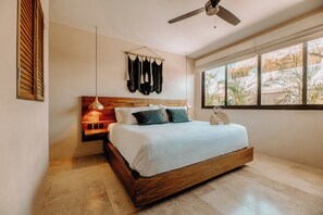 Une chambre moderne avec un lit en bois, un ventilateur de plafond, une grande fenêtre donnant sur des palmiers et une décoration murale en macramé.
