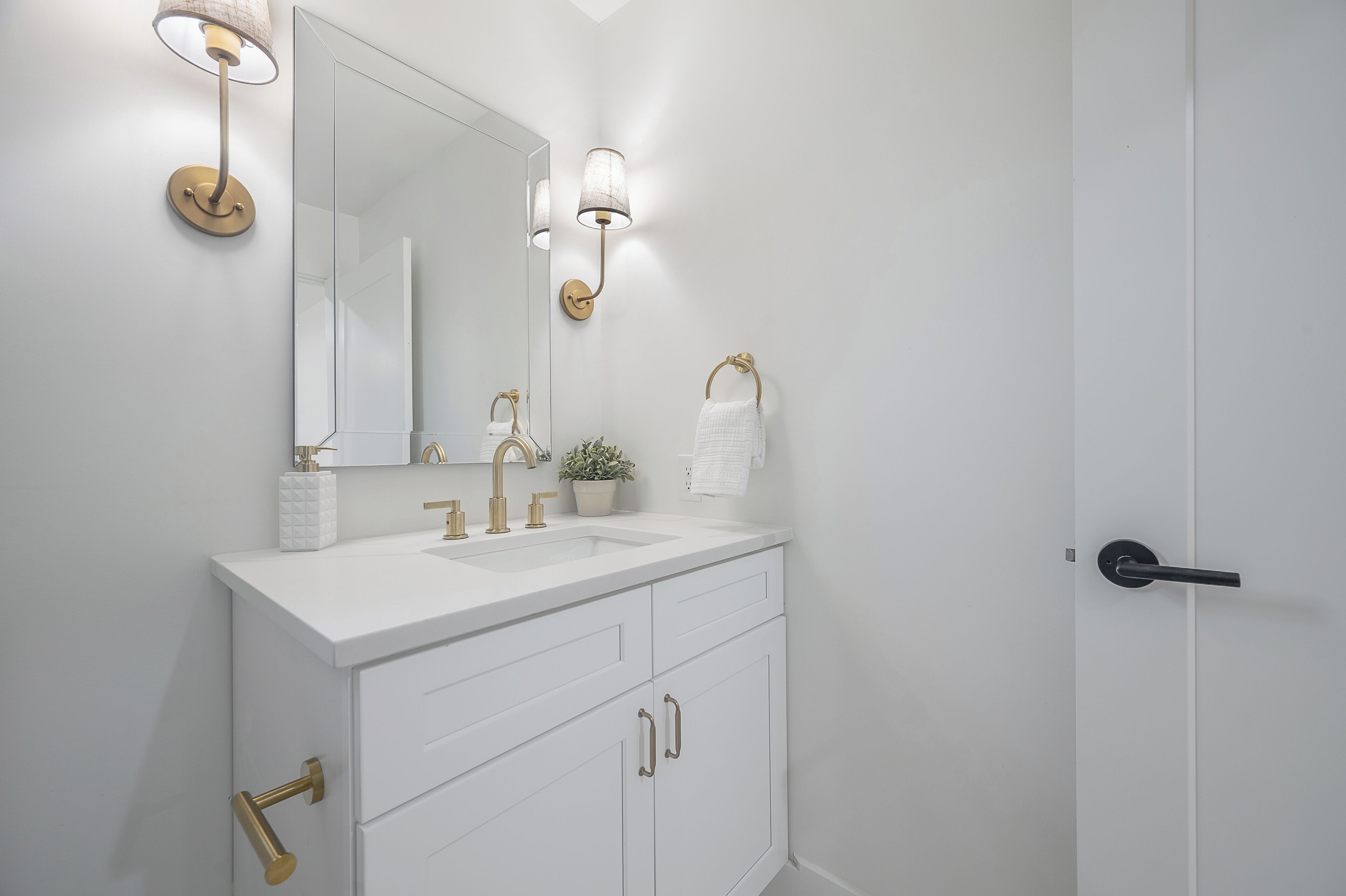 A white bathroom with a large mirror, gold fixtures, and a potted plant.