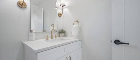A white bathroom with a large mirror, gold fixtures, and a potted plant.