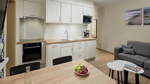 A modern kitchen with white cabinets, a microwave, and a sink. There is a dining table with a bowl of fruit and a glass bottle. A gray sofa and a small white table with a magazine are in the living area.