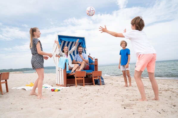 A beach scene with a beach chair, a person sitting in the chair, and another person standing nearby. - Appartements im Freizeitpark Weissenhauser Strand (Wangels)