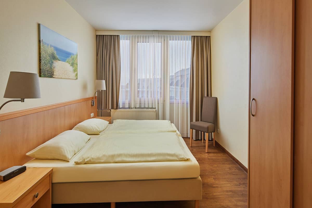A hotel room with a bed, a chair, a desk with a lamp, a painting on the wall, and a window with curtains.
