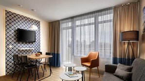 A modern hotel room with a wooden floor, a grey sofa, a round dining table with chairs, a small white table, a TV mounted on the wall, and a large window with curtains.