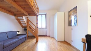 Ein Zimmer mit einer Holztreppe, einem grauen Sofa, einem Heizkörper, einem Fenster und einem Gemälde an der Wand.