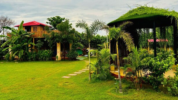 A lush garden with a pathway leading to a wooden structure with a red roof. - Aranyaa Cottage Homestay, Vrindavan (Jait)