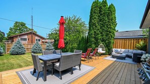 A deck with a dining table and chairs, a red umbrella, and a hanging chair. - Niagara 3BR w/ Huge Deck, King Suite, EV Charger & 5-Car Parking (St. Catharines)