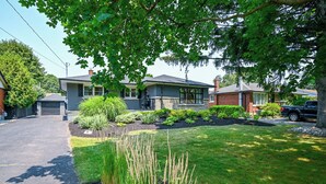 A residential street with houses, a driveway, and a well-maintained lawn. - Niagara 3BR w/ Huge Deck, King Suite, EV Charger & 5-Car Parking (St. Catharines)