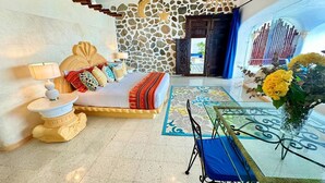 Habitación de hotel con pared de piedra, cama con almohadas coloridas, mesita de noche, una mesa con un jarrón de flores y vista al mar a través de una puerta de vidrio.