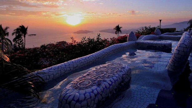 Un jacuzzi con suelo de mosaicos, rodeado de plantas tropicales y con vistas a un atardecer.