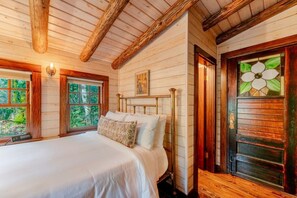 Quarto de cabana de madeira com uma cama, duas janelas, uma porta com design em vitral e teto de madeira.