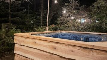 A wooden hot tub with a built-in seat, surrounded by trees and string lights.