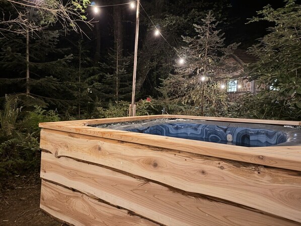 A wooden hot tub with a built-in seat, surrounded by trees and string lights.