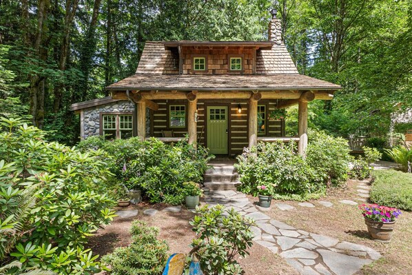 A rustic log cabin with a stone chimney, surrounded by lush greenery and a stone pathway leading to the entrance. - Creekside Fairytale Cabin Near Ferries to Seattle (Port Orchard)