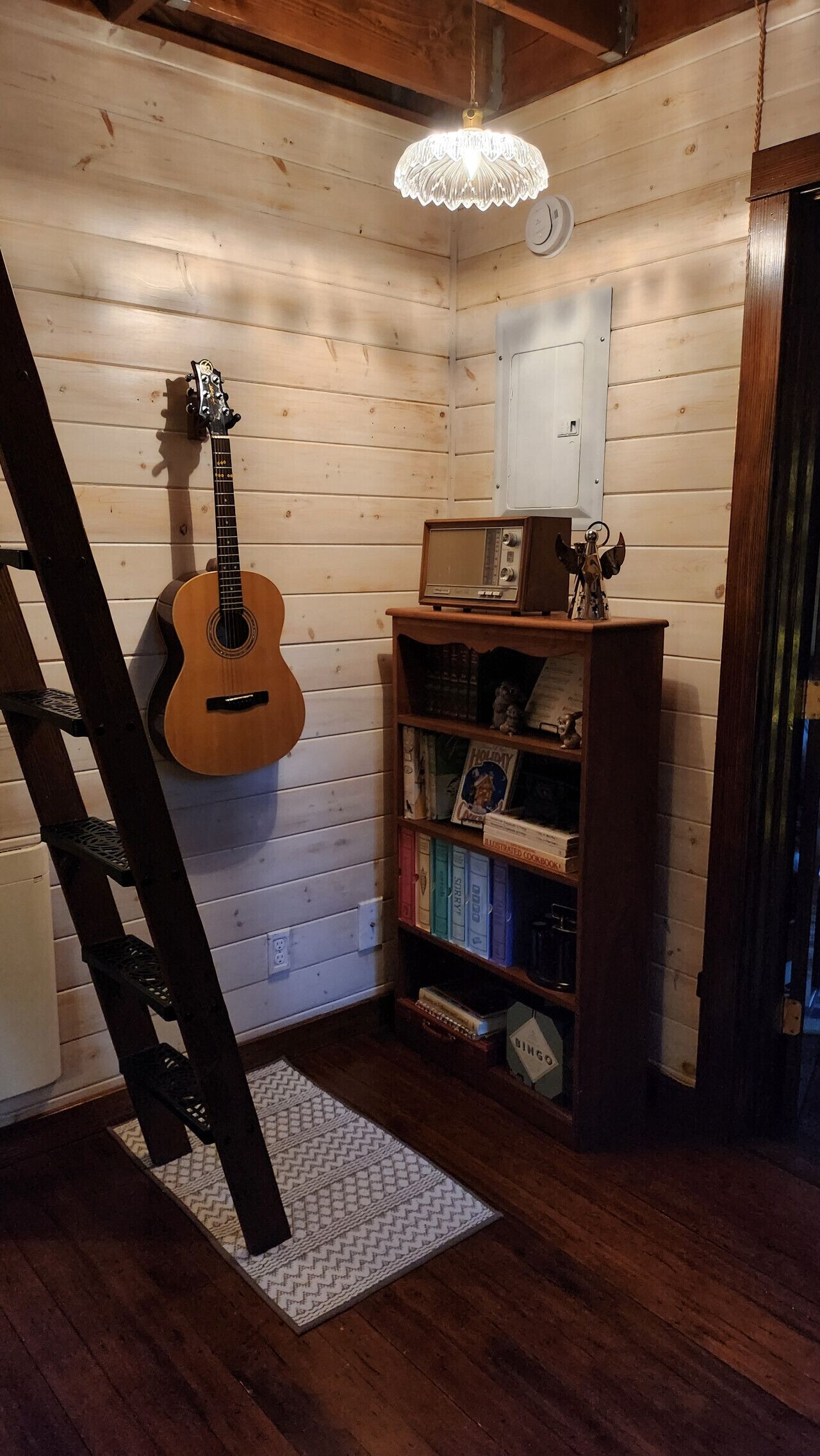 A wooden interior with a guitar leaning against the wall, a bookshelf with books and a radio, and a ladder.