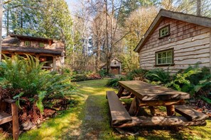 A rustic cabin with a wooden deck and a picnic table, surrounded by lush greenery and trees. - Creekside Fairytale Cabin Near Ferries to Seattle (Port Orchard)