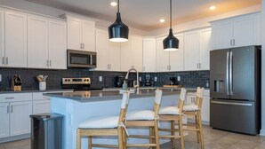 Una cucina moderna con mobili bianchi, un'isola centrale con sgabelli da bar, elettrodomestici in acciaio inossidabile e illuminazione a sospensione.