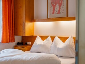 Quarto de hotel com cama, almofadas brancas, cabeceira de madeira, mesa de cabeceira com relógio e um quadro na parede.