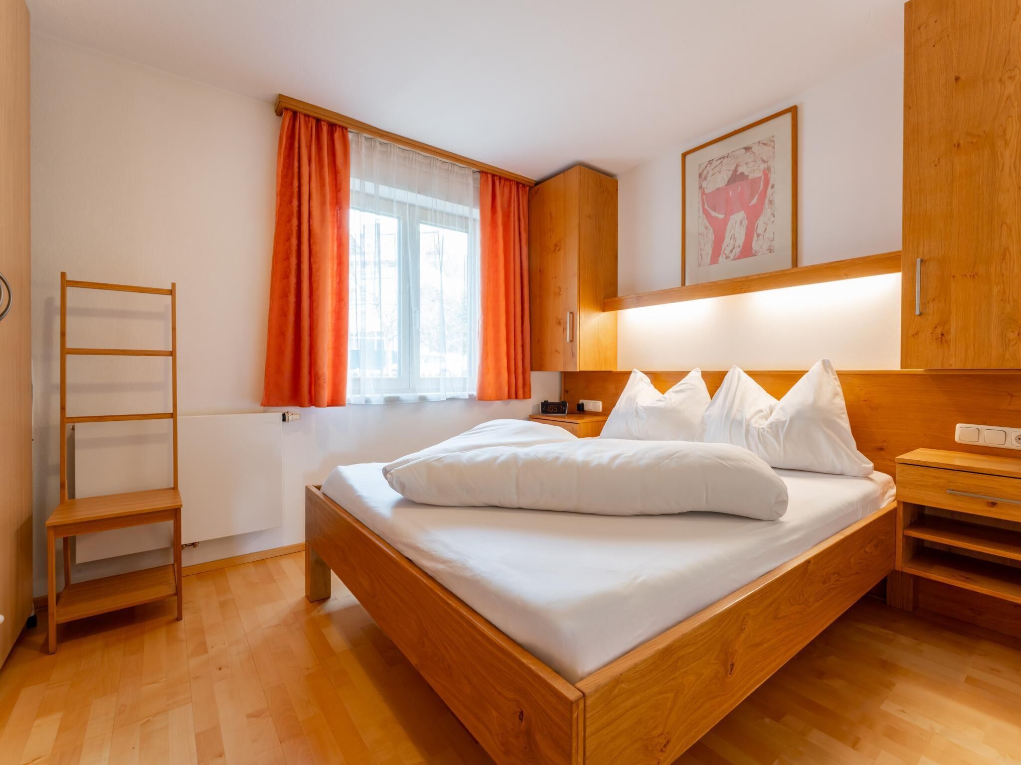 Un lit en bois avec une literie blanche, une table de chevet, une commode, une étagère en bois, une fenêtre avec des rideaux orange et un luminaire fixé au mur.