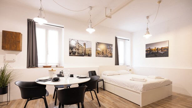 Moderni hotellihuone, jossa on sänky, ruokapöytä tuoleineen, ruukkukasvi ja taideteoksia seinillä.