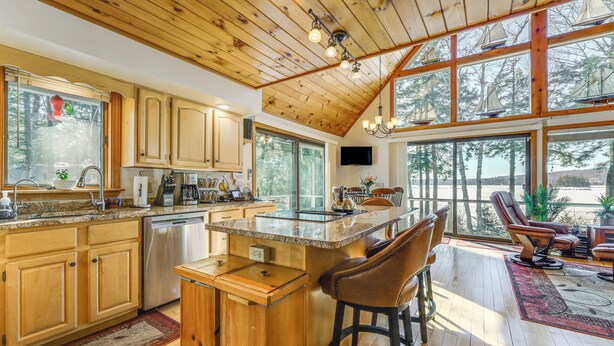 A spacious kitchen with wooden cabinets, a large island with bar stools, and a dining area with a view of the outdoors. - Gateway to Acadia! Green Lake Home w/ Hot Tub (Dedham)