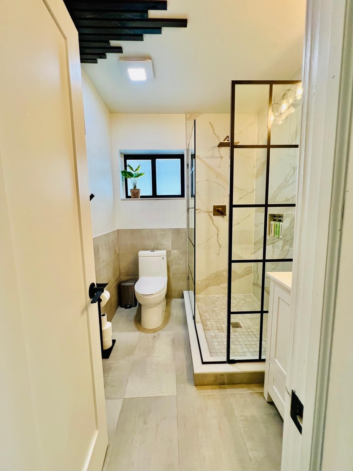 Et moderne badeværelse med toilet, glasvægs brusekabine og et lille vindue med en plante.