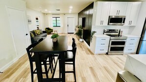 Una cocina moderna con gabinetes blancos, electrodomésticos de acero inoxidable y un comedor con mesa y sillas negras.