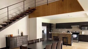 A modern kitchen with dark cabinetry, a central island, and a dining area with a large table and chairs. - Exquisite 3-BR Condo in the heart  of everything! (San Rafael de Escazú)