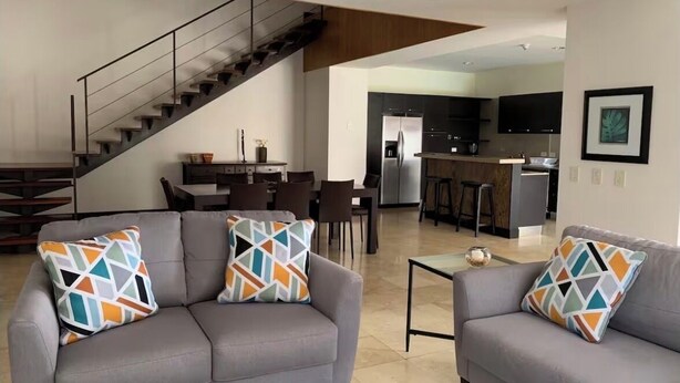 A modern living room with two grey sofas, a glass coffee table, and a staircase leading to a kitchen area. - Exquisite 3-BR Condo in the heart  of everything! (San Rafael de Escazú)