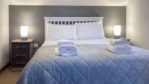 Een net opgemaakt bed met een blauw sprei, twee witte kussens en twee opgerolde handdoeken. Aan beide zijden van het bed staan nachtkastjes met lampen.
