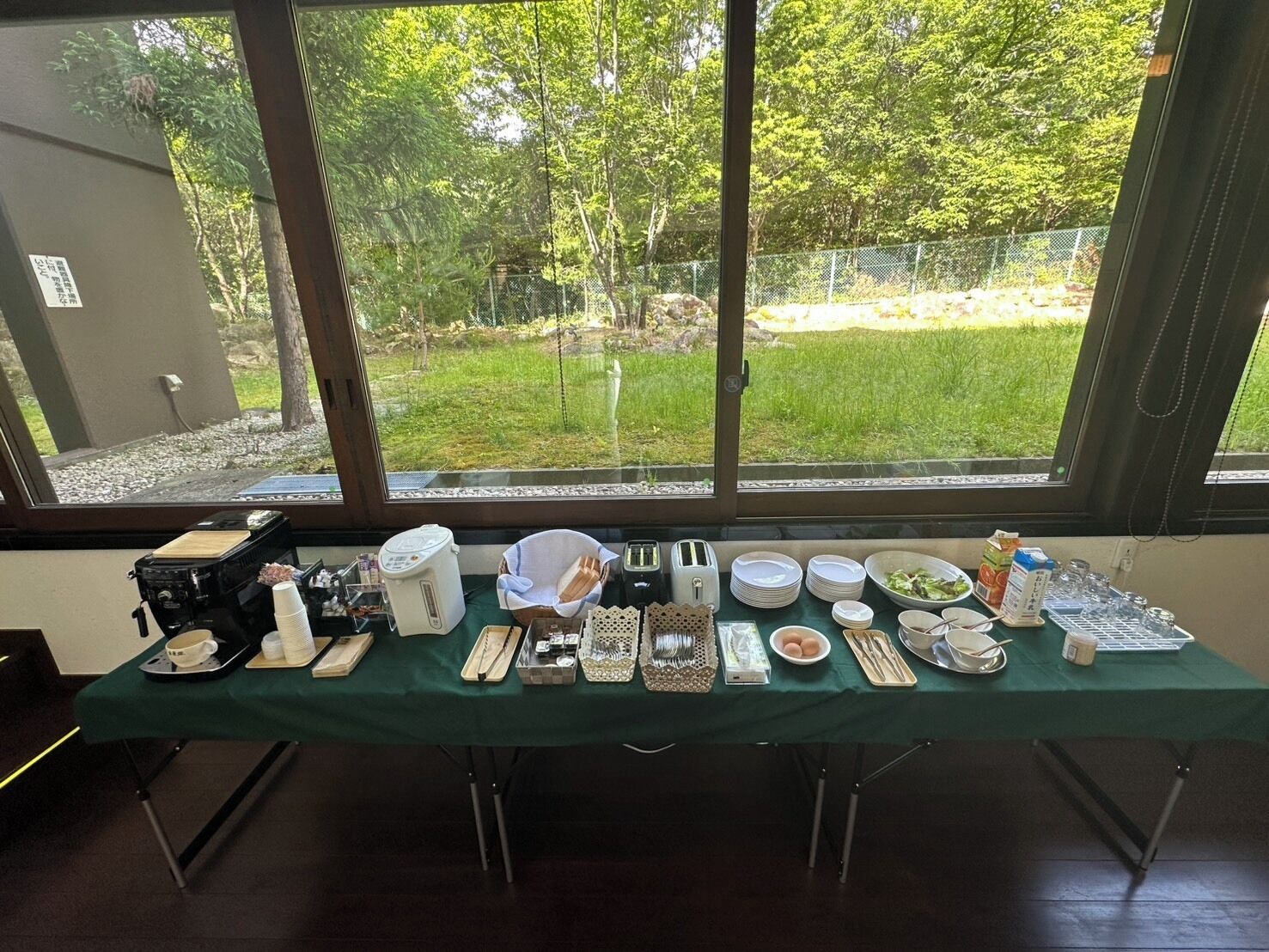 A table set with various dishes and utensils, a portable coffee maker, and a chair, with a view of a grassy area and trees through large windows.