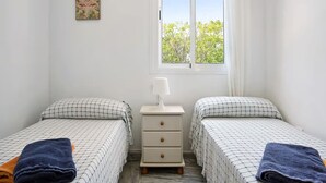 Ein kleiner, heller Raum mit zwei Betten, einem Nachttisch, einem Fenster mit Blick auf Bäume und einer Wanddekoration im tropischen Stil.